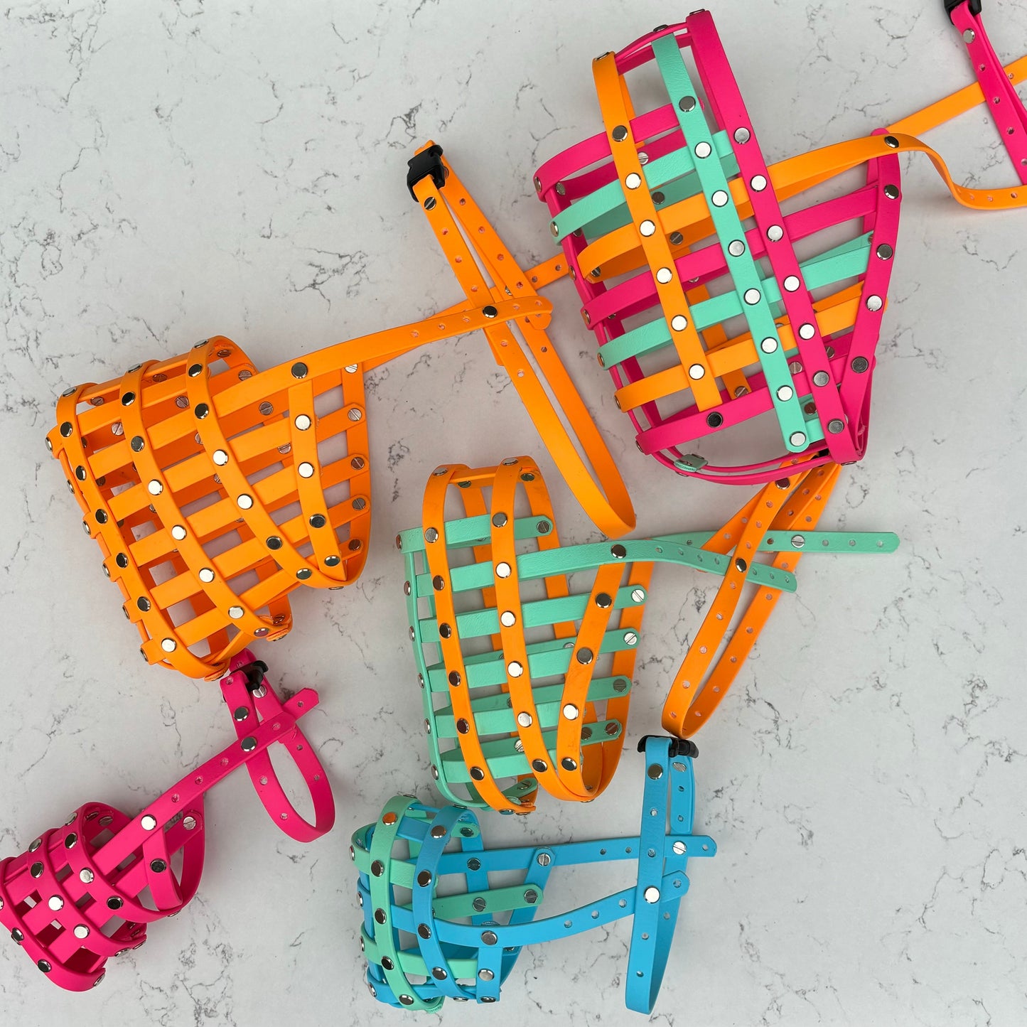A selection of custom handcrafted dog muzzles laid out on a surface, featuring various bright colors such as pink, orange, green, and blue. Each muzzle has a geometric pattern and includes a chin strap.